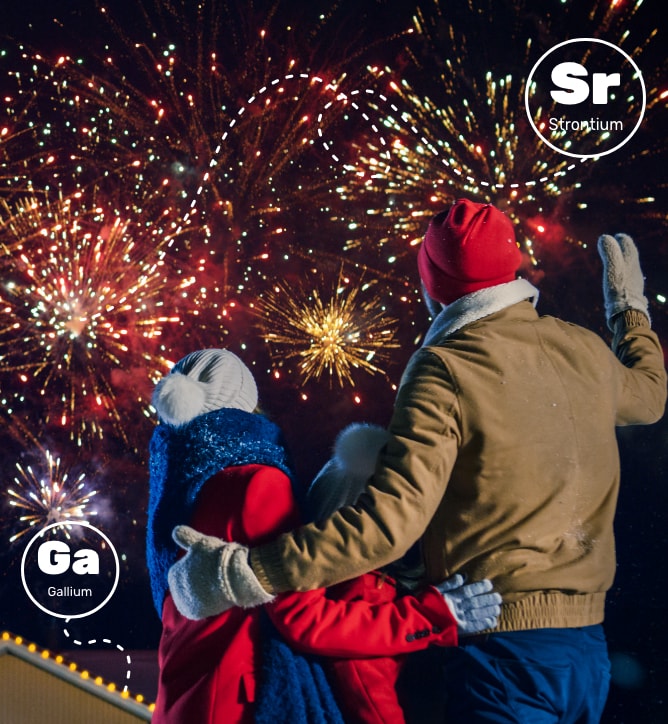 Image of a family enjoying fireworks – their backs are to the viewer and the fireworks are in the background above their house in the night sky. The edge of the roof is decorated with little lights. The family consists of a man, a woman, and a child. Strontium is used to create red fireworks and gallium is used in LED lights.