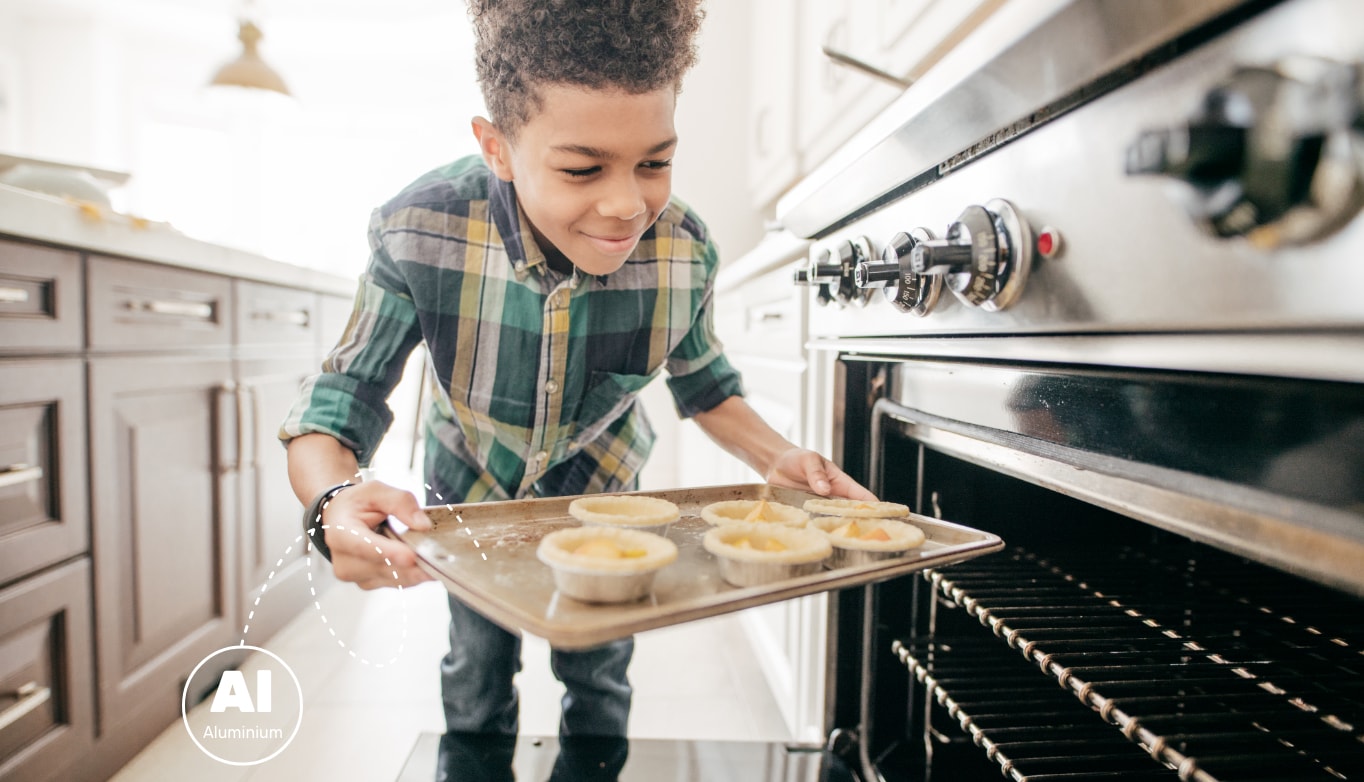 Image d'un enfant dans la cuisine en train de cuisiner. Il dépose des tartes dans un four ouvert. L'aluminium est souvent utilisé dans les ustensiles de cuisine, notamment les moules à pâtisserie.